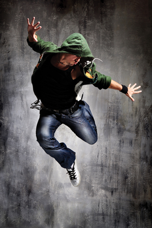 The photograph shows a male dancer wearing a green jacket and blue jeans leaping into the air.