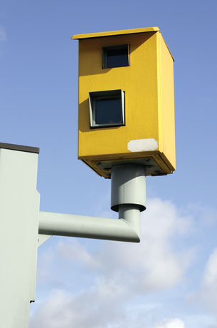 A red-light camera is perched on a pole in this photograph. The camera is housed in a yellow box.