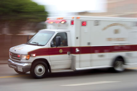 A photo shows an ambulance, with emergency lights turned on, driving fast on a road.
