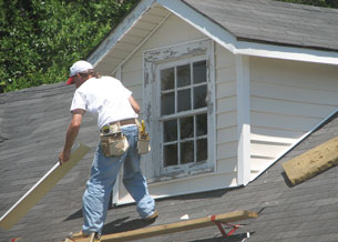 A photo shows a male roofer working on the roof of a house. He is standing on an incline. He has his foot placed on a board to prevent him from falling off the roof.