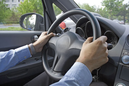 A photo shows a man’s arms holding onto the steering wheel of a car.