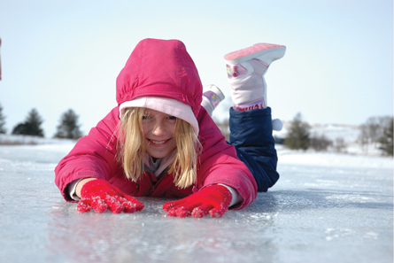 A young girl wearing a red parka and snow boots is playing on the ice in this photograph. She is smiling and laying on her stomach.