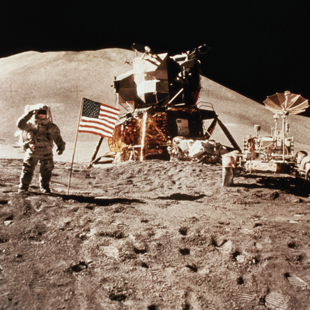 A photograph shows an astronaut standing next to a U.S. flag, a spacecraft, and a wheeled vehicle with assorted equipment.