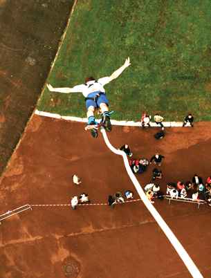 A photograph shows a jumper, who is attached to a bungee cord, jumping head first toward an open field below. There is a crowd of people watching from below.