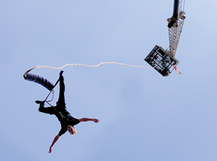 A photograph, aimed from the ground and pointing upwards, shows a male bungee jumper on his way down from a platform.