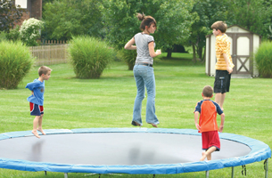 A photograph shows a woman jumping on a large, round trampoline. There are three young boys also on the trampoline.