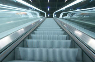 A photograph shows an escalator from the point of view of the bottom looking up.