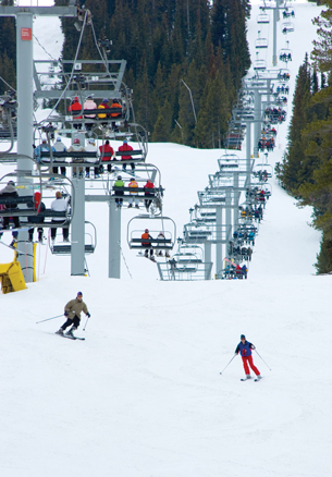 A photograph shows a series of chairs, all filled by skier, of a chairlift. There are also two skiers on the ground.