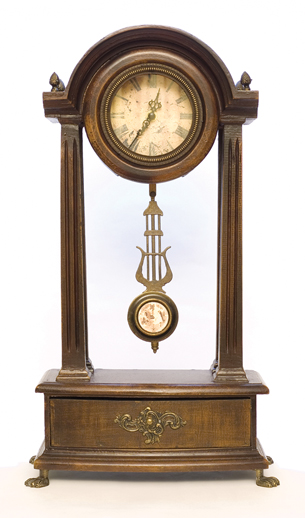 A photograph shows an ornamental clock with time piece at the top and held in place by two wooden posts that attach to a base at the bottom. Hanging from the time piece is a pendulum.