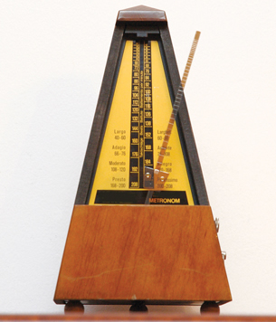 A metronome is shown in this photograph. It is shaped like a triangle—larger at the base and smaller at the top. There is vertical bar that is attached to the base but free at the top. A small weight is attached to the vertical bar.