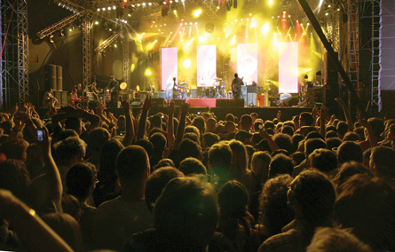 A large standing crowd is watching a group of musicians on a lighted stage in this photograph.